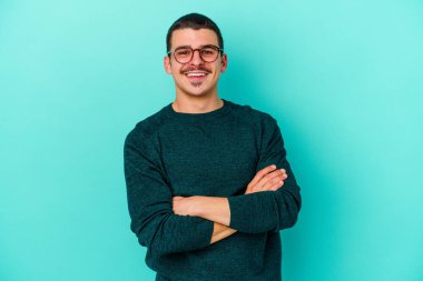 Mavi arka planda kendini güvende hisseden, kararlılıkla kollarını kavuşturan genç beyaz bir adam..