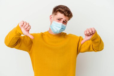 Koronavirüs maskesi takan genç adam beyaz arka planda izole edilmiş baş parmağını gösteriyor ve hoşlanmadığını ifade ediyor..