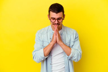 Sarı arka planda izole edilmiş, ağzı yakınken el ele dua eden, kendinden emin genç beyaz bir adam..