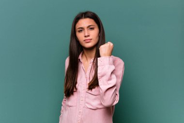 Mavi arka planda izole edilmiş genç Hintli kadın kameraya yumruk gösteriyor, agresif yüz ifadesi..