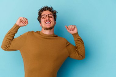 Mavi arka planda izole edilmiş gözlüklü genç beyaz adam zaferden sonra yumruğunu kaldırıyor, kazanan konsept..