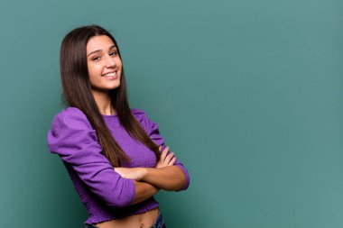 Mavi arka planda izole edilmiş genç Hintli kadın kollarını kavuşturarak gülümsüyor..