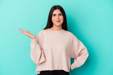 Kafkasyalı genç bir kadın mavi arka planda izole edilmiş sorgulama sırasında omuz silkiyor ve kuşkulanıyor..