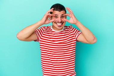 Mavi arka planda izole edilmiş genç beyaz adam bir başarı fırsatı bulmak için gözlerini açık tutuyor..