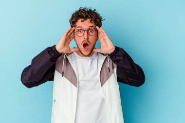Mavi arka planda gözlüklü genç beyaz adam yüzünün yarısını avuç içiyle kaplayarak eğleniyor..