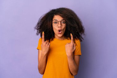 Genç Afro-Amerikalı kıvırcık kadın, mor arka planda izole edilmiş, ağzı açık, üst tarafı işaret ediyor..
