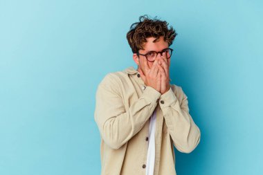Gözlüklü genç beyaz adam mavi arka planda izole edilmiş parmaklarıyla kameraya göz kırpıyor, yüzünü kapatmaktan utanıyor..