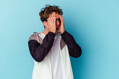 Mavi arka planda gözlük takan genç beyaz adam elleriyle gözlerini kapatmaktan korkuyor..