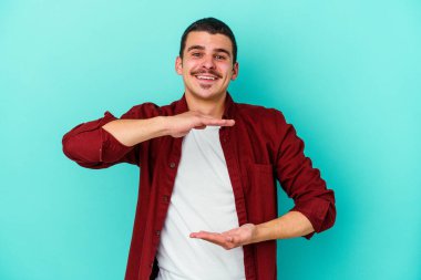 Mavi arka planda izole genç beyaz adam iki eliyle bir şey tutuyor, ürün sunumu..