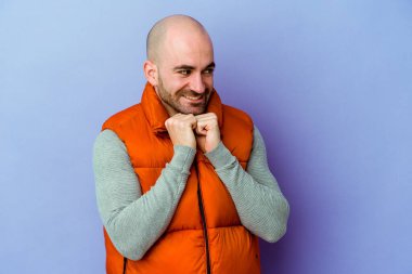 Mor arka planda izole genç beyaz kel adam ellerini çenesinin altında tutuyor, mutlu bir şekilde yana bakıyor..