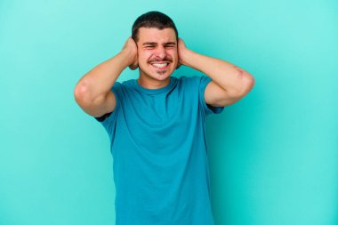 Mavi arka planda izole edilmiş genç beyaz adam kulaklarını elleriyle kapatıyor..