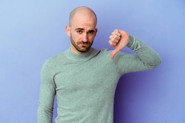 Mor arka planda izole edilmiş genç, beyaz, kel bir adam hoş olmayan bir jest yapıyor, başparmakları aşağıda. Anlaşmazlık konsepti.