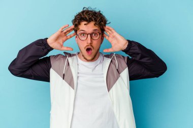 Mavi arka planda gözlüklü genç beyaz bir adam zaferi ya da başarıyı kutluyor, şaşırıyor ve şok oluyor..