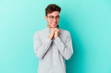 Mavi arka planda izole edilmiş genç beyaz adam ellerini çenesinin altında tutuyor, mutlu bir şekilde yana bakıyor..