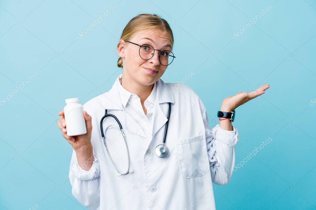 Young russian doctor woman holding pills bottle on blue doubting and shrugging shoulders in questioning gesture.