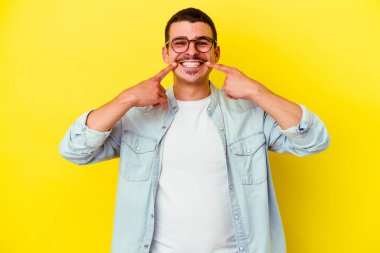 Sarı arka planda izole edilmiş genç beyaz adam gülümsüyor, parmaklarını ağıza doğrultuyor..