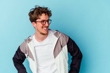 Mavi arka planda gözlük takan genç beyaz adam gülüyor ve gözlerini kapıyor, rahatlamış ve mutlu hissediyor..