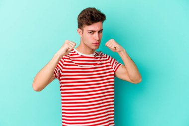 Kafkasyalı genç bir adam mavi arka planda izole edilmiş. Yumruk atıyor, sinirleniyor, kavga ediyor, kavga ediyor..