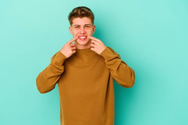 Mavi arka planda izole edilmiş genç beyaz adam gülümsüyor, parmaklarını ağıza doğrultuyor..