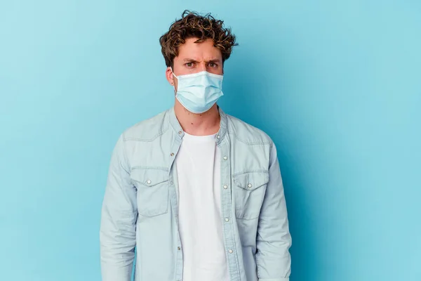Young caucasian man wearing an antiviral mask isolated on blue background blows cheeks, has tired expression. Facial expression concept.