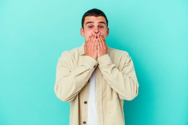 Mavi arka planda izole edilmiş genç beyaz adam şok olmuş, ağzı ellerle kaplanmış, yeni bir şey keşfetmeye hevesli..