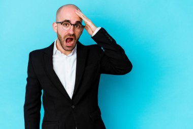 Genç, beyaz, kel bir adam mavi arka planda izole edilmiş, uzaklara bakıyor ve elini alnına koyuyor..