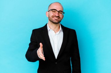 Genç, beyaz, kel bir adam mavi arka planda izole edilmiş. Kameraya doğru elini uzatarak selam veriyor..