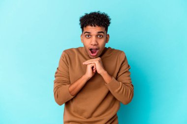 Genç Afro-Amerikalı kıvırcık adam mavi üzerine izole edilmiş şans için dua ediyor, şaşırmış ve ağzını açıyor..