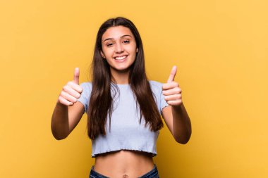 Sarı arka planda izole edilmiş genç Hintli kadın gülümsüyor ve başparmağını kaldırıyor.