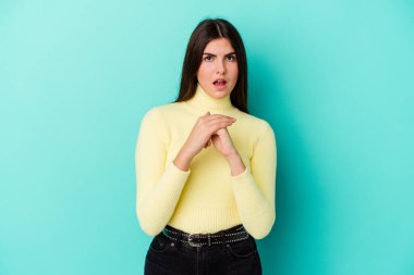 Kafkasyalı genç bir kadın mavi arka planda izole edilmiş şans için dua ediyor, şaşırmış ve ağzını açıp önüne bakıyor..