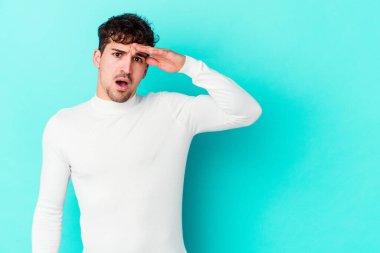 Kafkasyalı genç adam mavi arka planda izole edilmiş, uzaklara bakıyor ve elini alnına koyuyor..