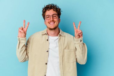 Mavi arka planda gözlüklü genç beyaz adam zafer işareti gösteriyor ve gülümsüyor..