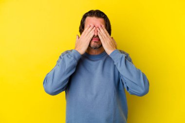Sarı arka planda izole edilmiş orta yaşlı beyaz bir adam elleriyle gözlerini kapatmaktan korkuyor..
