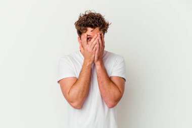 Beyaz arka planda izole edilmiş genç beyaz bir adam parmaklarının arasından korkmuş ve gergin bir şekilde göz kırpıyor..