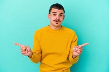 Mavi arka planda izole edilmiş genç beyaz adam farklı kopya alanlarını gösteriyor, birini seçiyor, parmaklarıyla gösteriyor.