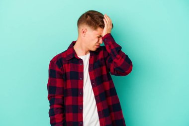 Kafkasyalı genç adam mavi arka planda izole edilmiş bir şekilde bir şeyi unutuyor. Alnına avuç içi ve gözlerini kapayarak vuruyor..