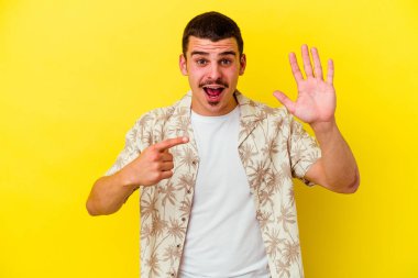 Sarı arka planda izole edilmiş genç beyaz adam gülümseyerek beş numarayı parmaklarıyla gösteriyor..