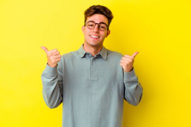Sarı arka planda izole edilmiş genç beyaz adam iki parmağını da kaldırıyor, gülümsüyor ve kendinden emin..