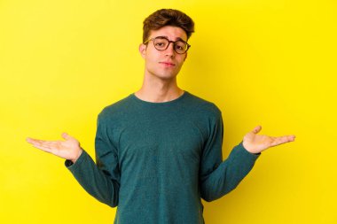 Sarı arka planda izole edilmiş genç beyaz adam sorgulama sırasında omuz silkiyor ve kuşkulanıyor..