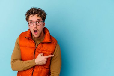 Mavi arka planda izole edilmiş gözlüklü genç beyaz adam gülümsüyor ve boş alanda bir şeyler gösteriyor..