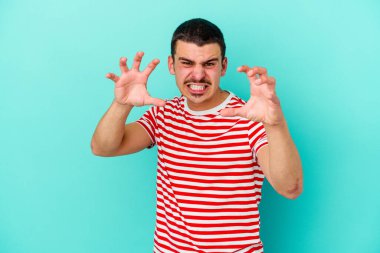 Mavi arka planda izole edilmiş genç beyaz adam pençelerini kedi taklidi yaparken gösteriyor, agresif bir hareket..