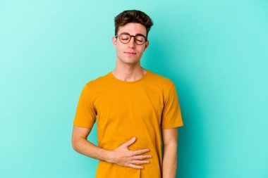 Mavi arka planda izole edilmiş genç beyaz adam karnına dokunuyor, nazikçe gülümsüyor, yemek yiyor ve tatmin oluyor..