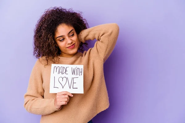 Young African American woman holding a Made with love placard isolated on purple background touching back of head, thinking and making a choice.