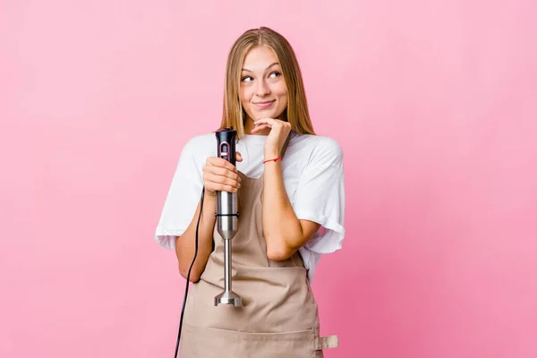 Young russian cook woman holding an electric mixer isolated keeps hands under chin, is looking happily aside.