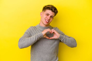 Sarı arka planda izole edilmiş genç beyaz adam gülümsüyor ve elleriyle kalp şeklini gösteriyor..