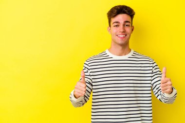 Sarı arka planda izole edilmiş genç beyaz adam gülümsüyor ve başparmağını kaldırıyor.