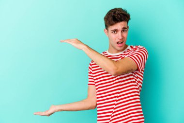 Mavi arka planda izole genç beyaz adam şok ve hayret içinde elleri arasında bir kopya boşluk tutuyor.