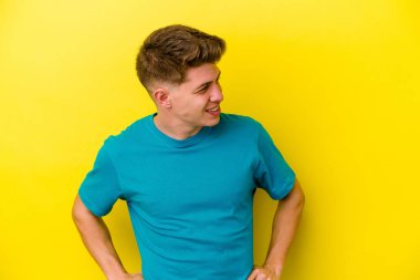 Sarı arka planda izole edilmiş genç beyaz adam mutlu bir şekilde gülüyor ve ellerini karnının üstünde tutarak eğleniyor..