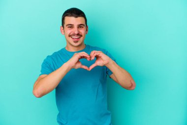 Mavi arka planda izole edilmiş genç beyaz adam gülümsüyor ve elleriyle kalp şeklini gösteriyor..