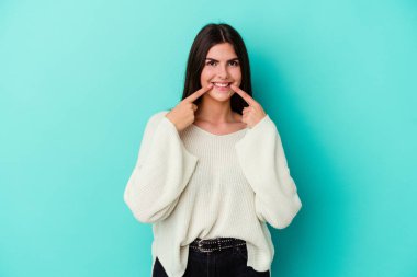 Mavi arka planda izole edilmiş genç beyaz kadın gülümsüyor, parmaklarını ağıza doğrultuyor..